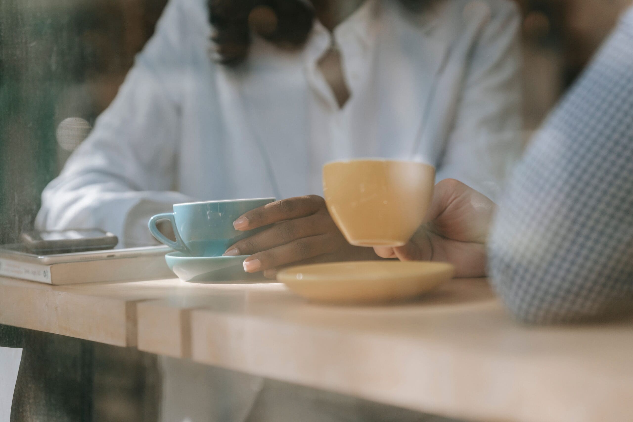 Two people sit at a café table by a window, each holding a coffee cup; their faces are out of frame.