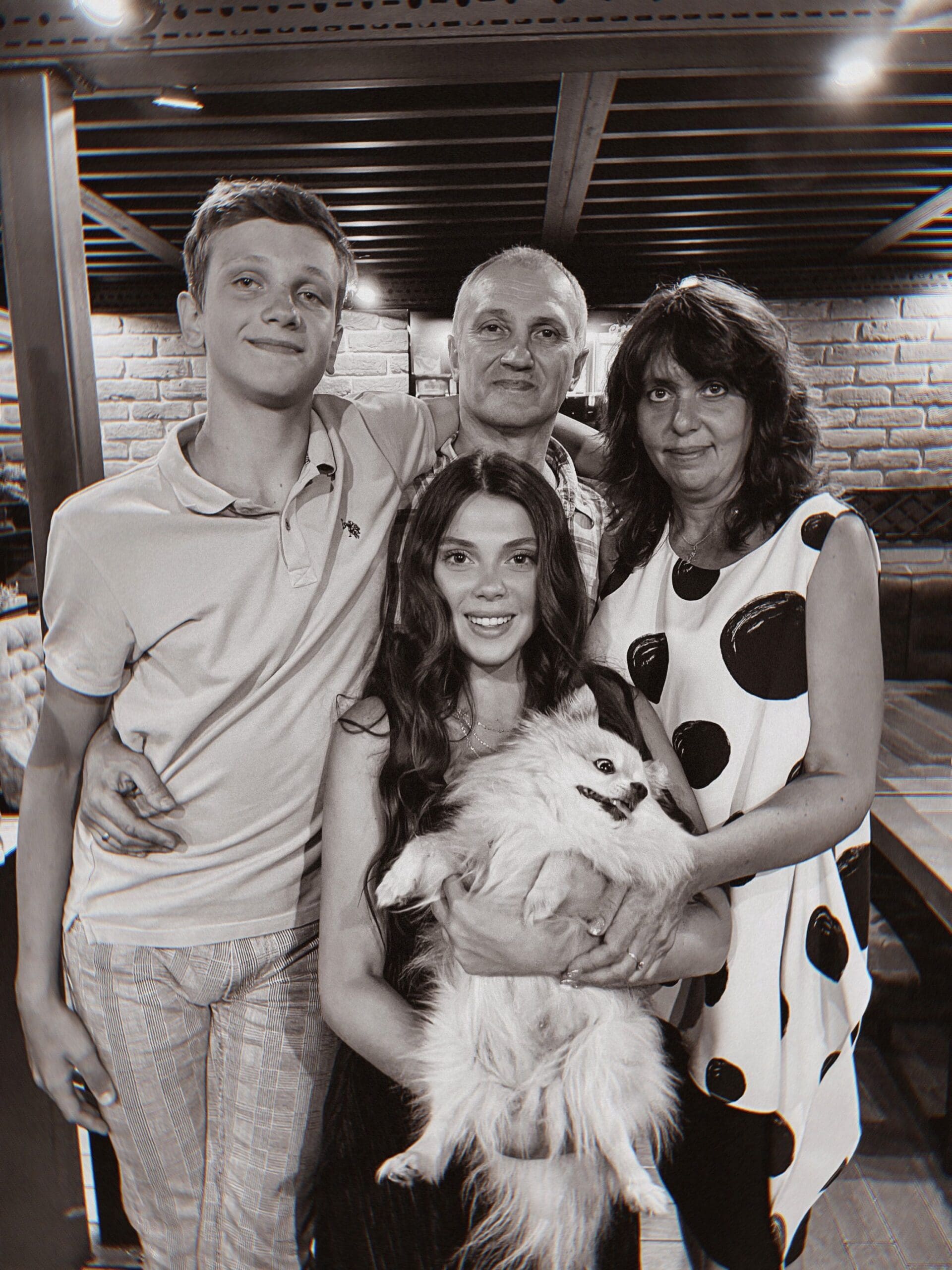 A black-and-white portrait of a family of four posing together indoors; a young woman at the center holds a small fluffy dog.
